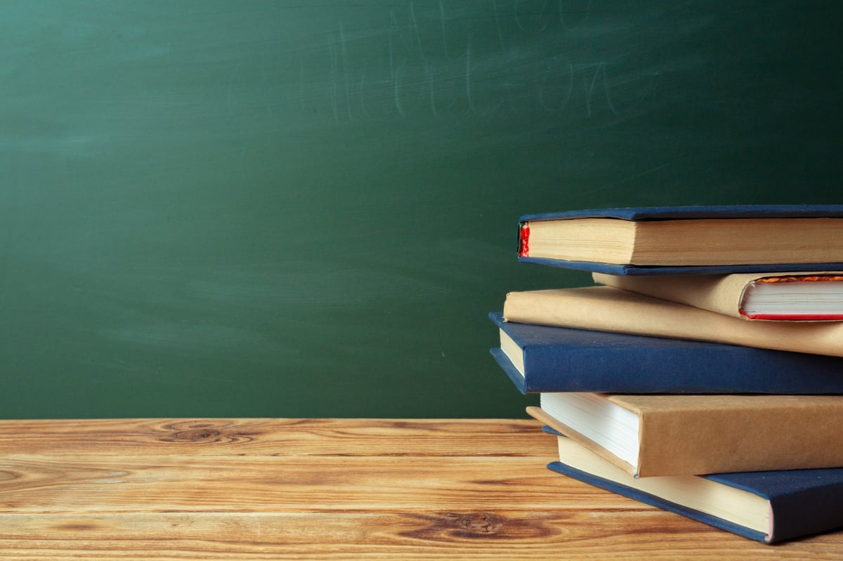 A collection of books is arranged on a wooden surface near a green chalkboard in a classroom.